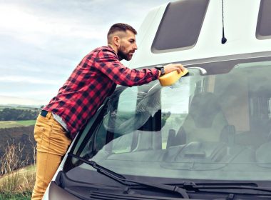 A rugged man in a plaid shirt leans over his camper van windshield and washes it with a sponge.