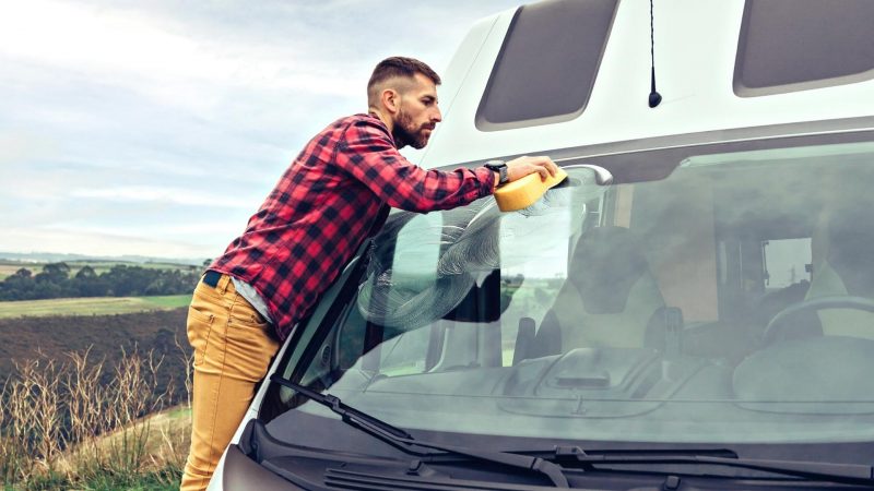 A rugged man in a plaid shirt leans over his camper van windshield and washes it with a sponge.