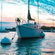 A sailboat anchored in a bay off the coast of a port city as the sun sets.