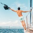 A man leans off the side of a sailboat as it cruises through the ocean on a beautifully sunny day.