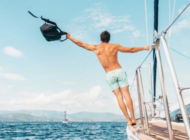 A man leans off the side of a sailboat as it cruises through the ocean on a beautifully sunny day.