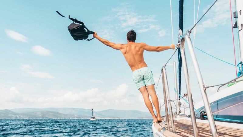 A man leans off the side of a sailboat as it cruises through the ocean on a beautifully sunny day.
