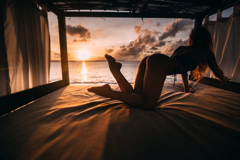 A woman in a swimsuit reaching for something on a bed in a luxurious bunk near the ocean sunset.