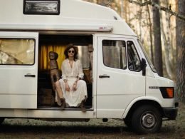 A woman slouches, seating in the sliding doorway of her white camper van in the woods.