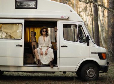 A woman slouches, seating in the sliding doorway of her white camper van in the woods.