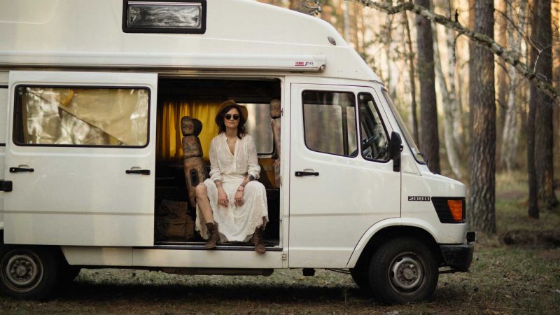 A woman slouches, seating in the sliding doorway of her white camper van in the woods.