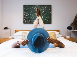 A man poses on a hotel bed with a big hat and feet up in the air.