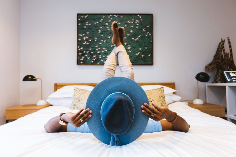 A man poses on a hotel bed with a big hat and feet up in the air.