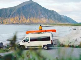 A woman sits on top of her camper van with a red canoe next to her overlooking a lake and a mountain.