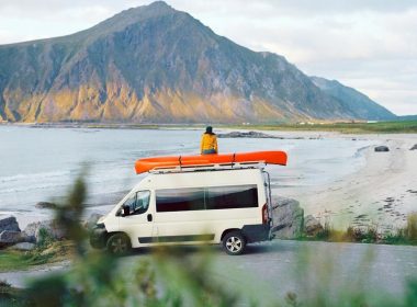 A woman sits on top of her camper van with a red canoe next to her overlooking a lake and a mountain.