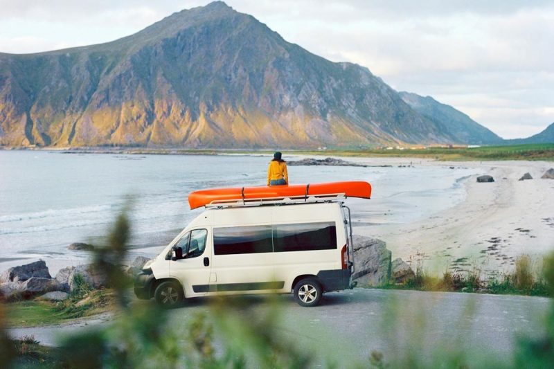 A woman sits on top of her camper van with a red canoe next to her overlooking a lake and a mountain.