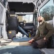 A man sits in his gutted sprinter van looking around and planning his camper build.