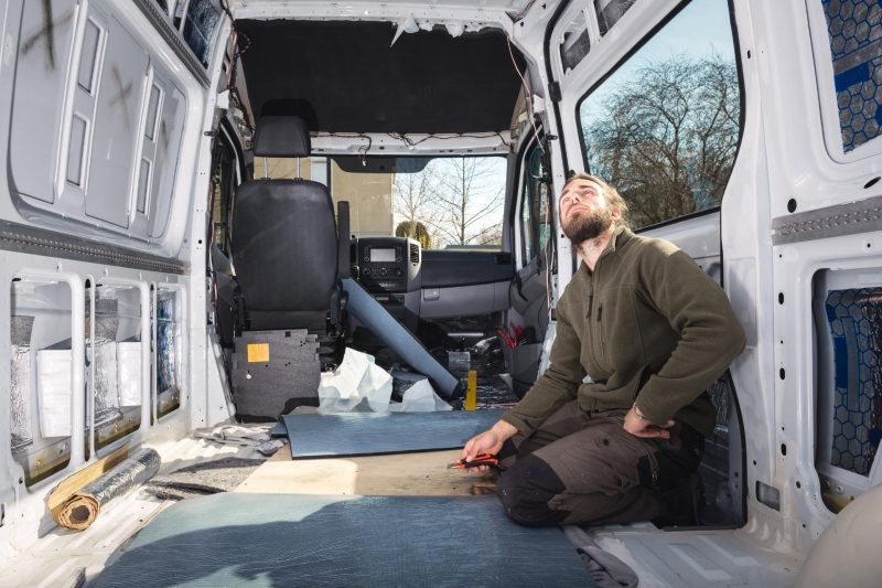 A man sits in his gutted sprinter van looking around and planning his camper build.