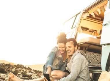 A couple sitting on the ground outside of their RV using a mobile phone.