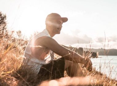 A man sits in the tall grass near the waterfront as the sun sets.