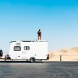 A woman stands on top of her travel trailer and looks around an empty parking lot in a remote desert.