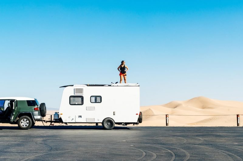 A woman stands on top of her travel trailer and looks around an empty parking lot in a remote desert.