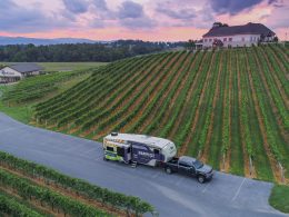 A travel trailer branded in the Harvest Host colors and logo parked at a beautiful winery at sunset.