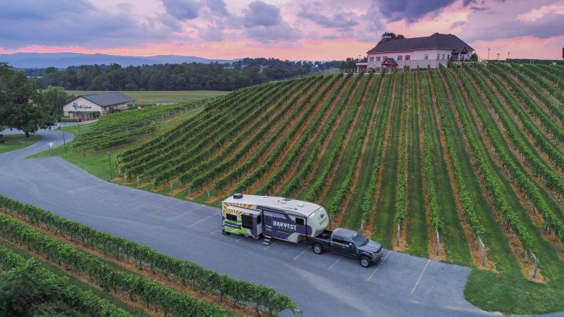 A travel trailer branded in the Harvest Host colors and logo parked at a beautiful winery at sunset.
