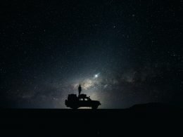 A night sky with stars and satellites illuminates the silhouette of a figure standing on top of an offroad vehicle looking up at the sky.