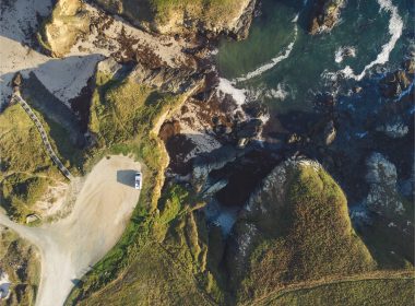An aeiral view of an RV parked on a cliff near water with waves crashing onto the rocky shore.