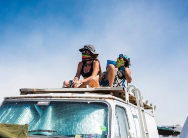 A couple takes a break on the roof of their van in the sun with bandanas over their faces.