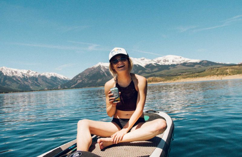 A woman sits on a paddle board in a lake drinking a beer from a can with snow capped mountains behind her.