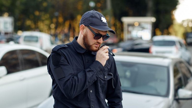 A police office talks into his radio on his shoulder while standing in a parking lot.