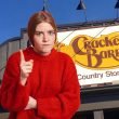 An upset woman waves a finger to reprimand in front of the Cracker Barrel building sign.
