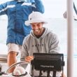 A happy young man smiles as he steers a sailboat out on the ocean with his buddy standing behind him.
