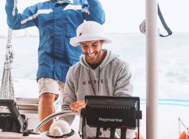 A happy young man smiles as he steers a sailboat out on the ocean with his buddy standing behind him.