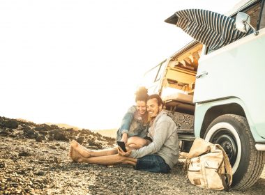 A couple sitting on the ground outside of their RV using a mobile phone.