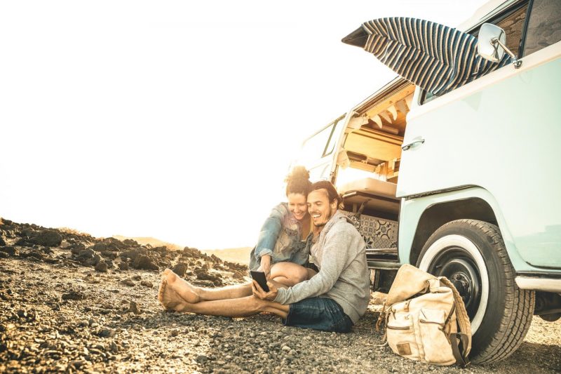 A couple sitting on the ground outside of their RV using a mobile phone.