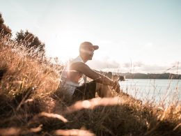 A man sits in the tall grass near the waterfront as the sun sets.