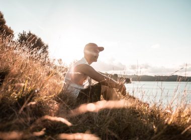 A man sits in the tall grass near the waterfront as the sun sets.