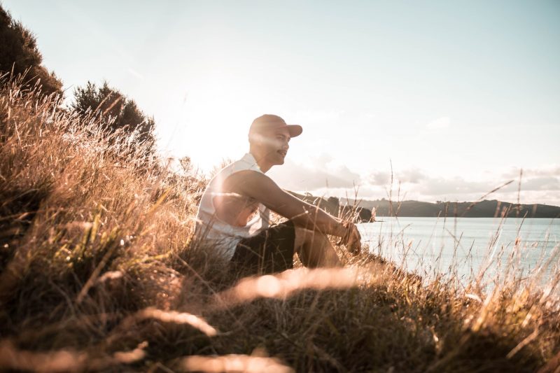 A man sits in the tall grass near the waterfront as the sun sets.