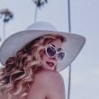 An actress with curly blond hair, sunglasses, and a large hat turns to look over her shoulder.
