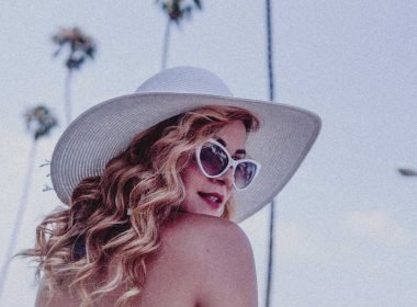 An actress with curly blond hair, sunglasses, and a large hat turns to look over her shoulder.