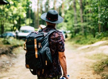A man wearing a hiking backpack looks on to a crowded campsite in the woods.