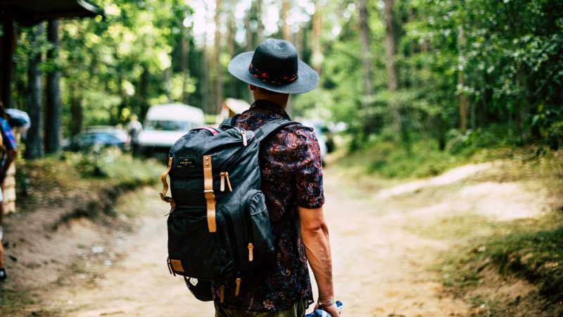 A man wearing a hiking backpack looks on to a crowded campsite in the woods.