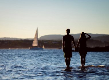 A dark silhoutte of a couple holding hands in the water with a sailboat in the distance.