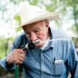 An older man with a white cowboy hat drinks from his garden hose outside.