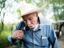 An older man with a white cowboy hat drinks from his garden hose outside.