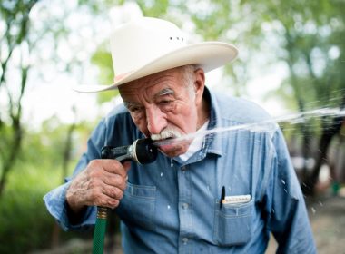 An older man with a white cowboy hat drinks from his garden hose outside.