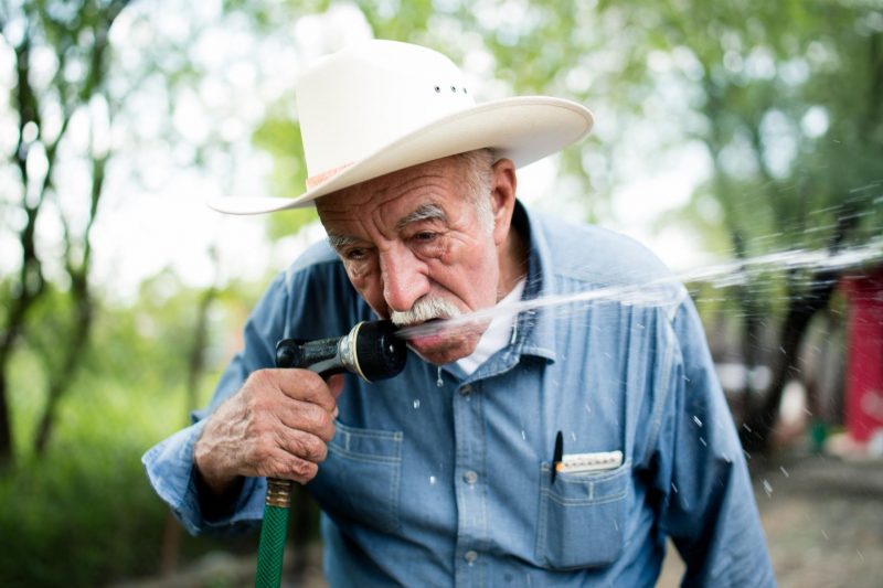 An older man with a white cowboy hat drinks from his garden hose outside.