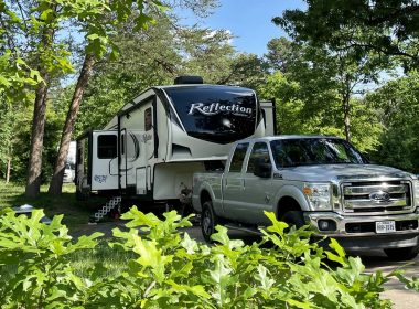A white truck towing a Grand Design Reflection fifth wheel through a beautiful summer campground.