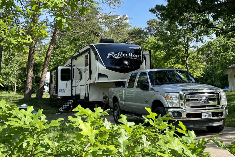 A white truck towing a Grand Design Reflection fifth wheel through a beautiful summer campground.