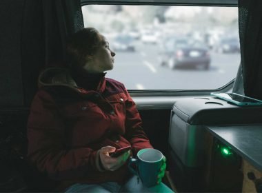 A woman drinking tea in her camper van looks out the window into a parking lot.