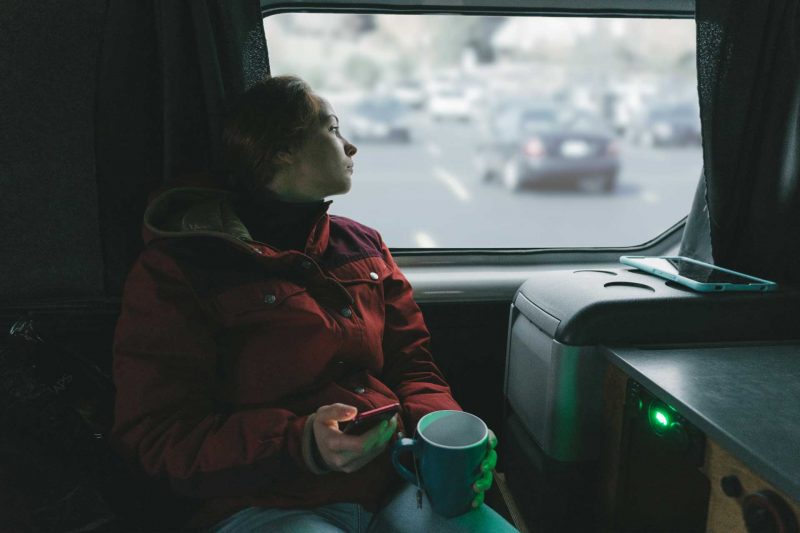 A woman drinking tea in her camper van looks out the window into a parking lot.