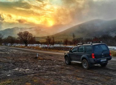 A Nissan Xterra parked on the side of a dirt road as the sun rises over foggy mountains.